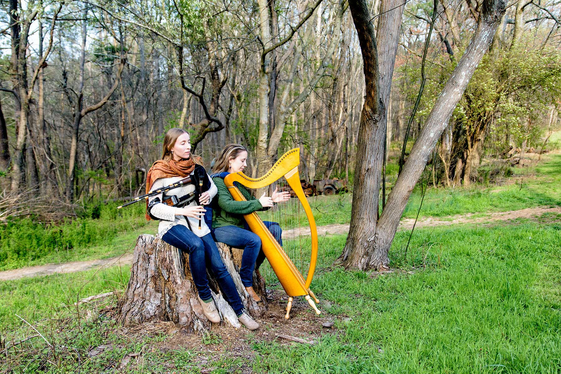 The Reel Sisters sitting on a tree stump. Rosalind is playing the Scottish smallpipes. Kelly is playing the harp.
